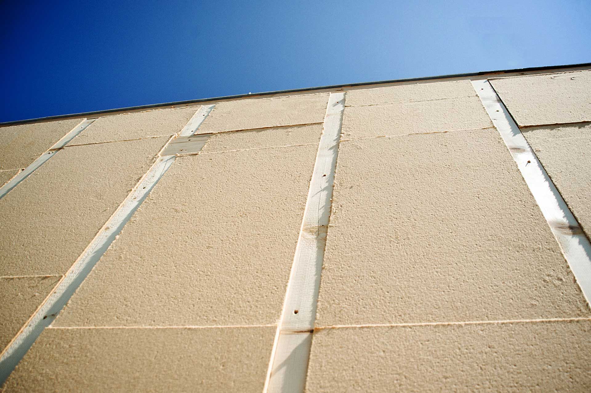 In primo piano si vedono pannelli isolanti in fibra di legno posati tra i listelli di una parete a telaio. Sullo sfondo, oltre la gronda, cielo azzurro.